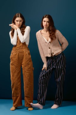 İkiz kız kardeşler etkileyici giysilerde eşsiz stiller sergilerler canlı mavi bir arka plana karşı.