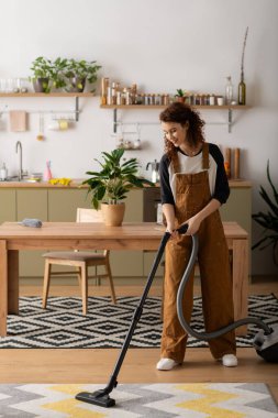 Kıvırcık saçlı genç bir kadın modern dairesini süpürürken gündelik kıyafetler giyer ve gülümser..