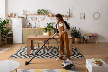 Kıvırcık saçlı, günlük kıyafetler giyen genç bir kadın şık dairesini süpürgeyle temizliyor..