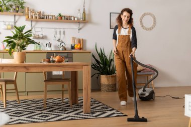 Kıvırcık saçlı bir kadın günlük giysiler içinde şık dairesini sıcak ve özenle temizliyor..