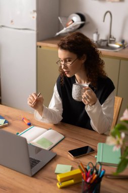 Kıvırcık saçlı genç bir kadın dairesinde renkli notlarla çevrili bir şekilde ders çalışıyor..