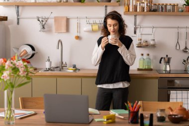 Kıvırcık saçlı genç bir kadın yaratıcı çalışma alanında dinlenirken çayını yudumluyor..