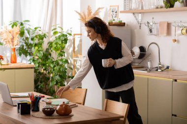 Kıvırcık saçlı genç bir kadın çalışma alanını şık bir dairede düzenliyor, kahvesini yudumluyor..