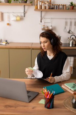 Kıvırcık saçlı genç bir kadın, kitaplarla çevrili, rahat bir dairede derslerine odaklanıyor..