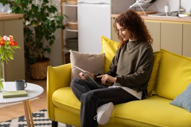 Kıvırcık saçlı kadın rahat dairesinde tablet kullanırken parlak bir kanepede dinleniyor..