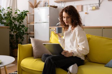 Şık bir dairede kıvırcık saçlı genç bir kadın canlı bir kanepede dinleniyor ve çayını yudumluyor..