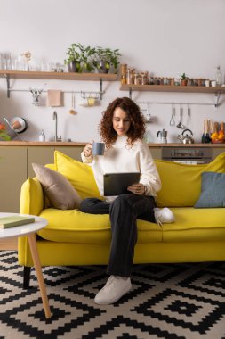 Modern bir dairede, kıvırcık bir genç kadın elinde içki tutarken parlak sarı bir kanepede dinlenir..