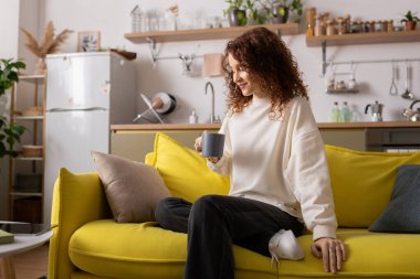 Genç bir kadın parlak sarı bir kanepede rahat rahat çayını yudumluyor..