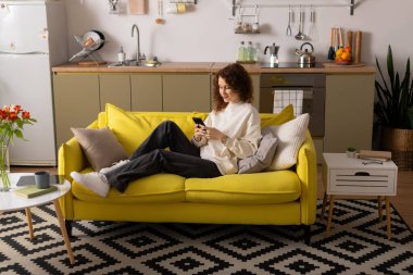 Gündelik giysiler içindeki kıvırcık bir kadın telefonuyla güneşli sarı bir kanepede vakit geçirmekten zevk alıyor..