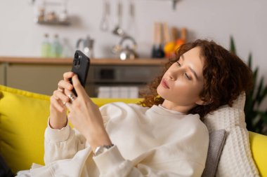 Genç bir kadın evde vakit geçirmeyi seviyor, rahat bir dairede akıllı telefonuyla kayıyor..