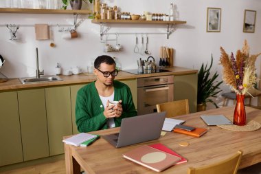 Kendine güveni tam genç bir adam, sıcak bir içecek ve rahat bir evde dizüstü bilgisayarla güzel bir masada dinleniyor..