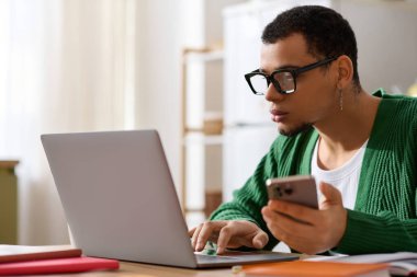 Sıcak ve davetkar bir ortamda, şık bir genç adam elinde telefon tutarken dizüstü bilgisayarına odaklanır..