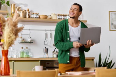 Gülümseyen genç adam sıcak bir mutfakta elinde dizüstü bilgisayarla evde eğleniyor..