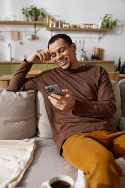 Yakışıklı genç adam kanepede rahat bir şekilde uzanıyor, akıllı telefonunu kullanarak gülümsüyor..