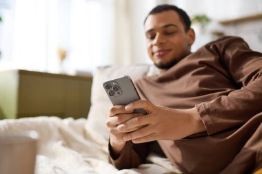 Genç bir adam, akıllı telefonuyla iletişim kurarken gülümseyerek evinde vakit geçirmekten zevk alır..