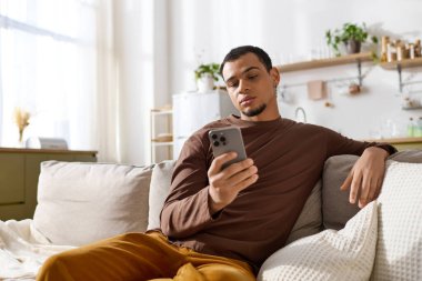 Young man lounges comfortably on a sofa, focused on his smartphone in a serene home setting.