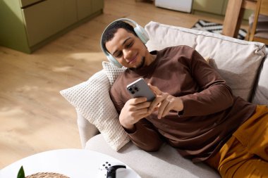 Genç adam kulaklık takıyor, gülümsüyor ve rahat bir kanepede uzanırken akıllı telefonunu kullanıyor..
