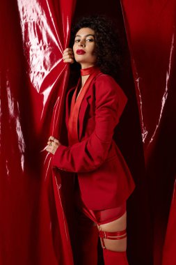 Kıvırcık saçlı kadın, göz alıcı bir kırmızı elbise sergiliyor, canlı bir stüdyo ortamında aşkı kutluyor..