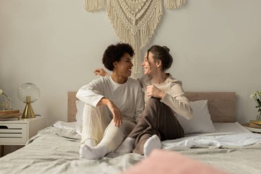 İki kadın yumuşak bir yatakta birlikte oturur, gülümser ve hoş bir karşılama odasında hassas bir anın tadını çıkarırlar..