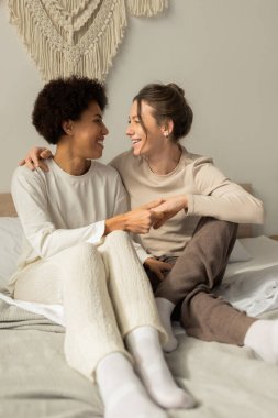 İki kadın yumuşak bir yatakta yan yana oturur, gülümser ve el ele tutuşur, sevgi ve mutluluk saçar..