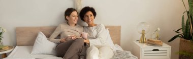 Two women snuggle on a bed, enjoying warm drinks and each others company in a sunny room.
