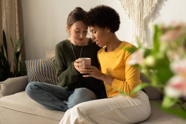 İki kadın birbirine yakın oturuyor, şefkatli gülümsemeler sergiliyor ve sıcak bir fincan kahve içiyorlar..