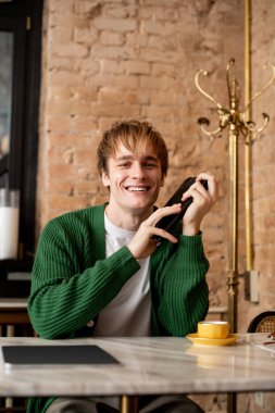 Sarışın genç adam telefonunu tutarken gülümsüyor, kahve molası veriyor..