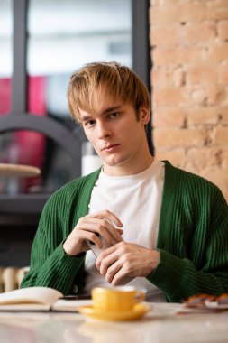 Sarı saçlı genç bir adam şık bir kafede rahatlar, kahvesini yudumlar ve yansıtır..