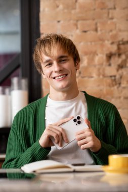 Sarışın genç adam rahat bir kafede telefonunu tutarken gülümsüyor..