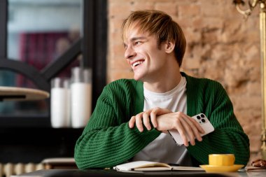 Genç sarışın bir adam modern bir kafede oturmuş hayattan zevk alırken rahatlıyor ve gülümsüyor..