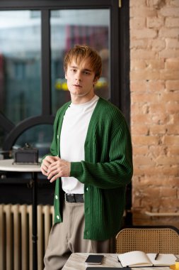 Sarışın genç adam, şık bir kafede sıcaklıkla çevrili dururken düşünceli görünüyor..