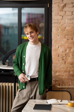 Sarı saçlı genç bir adam modaya uygun bir kafede durup, telefonunu tutuyor ve gülümsüyor..