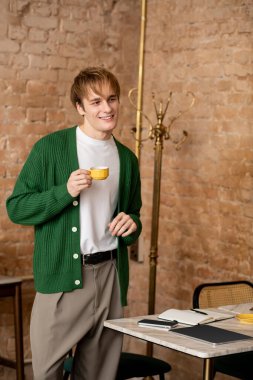 Havalı giyinmiş sarışın genç adam şık bir kafede elinde bir bardak tutarken gülümsüyor..