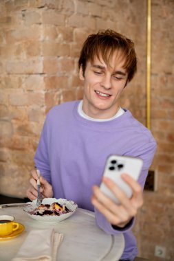 Sarı saçlı genç adam şık bir kafe ortamında mesajlaşırken tatlının tadını çıkarıyor..