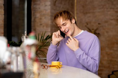 Sarı saçlı genç bir adam şık bir kafede telefonla konuşurken gülüyor..