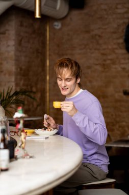 Sarı saçlı neşeli genç bir adam modern bir kafede lezzetli bir yemeğin tadını çıkarıyor..