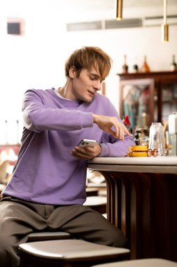 Havalı kıyafetler içindeki sarışın genç adam şık bir kafede telefonunu kullanırken gülümsüyor..