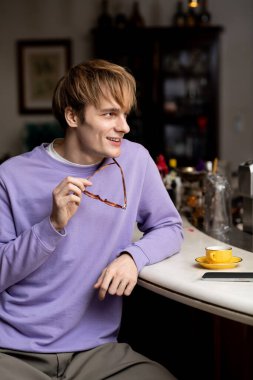 Sarı saçlı genç bir adam şık bir kafede dinlenirken gülümsüyor, gözlük takıyor..