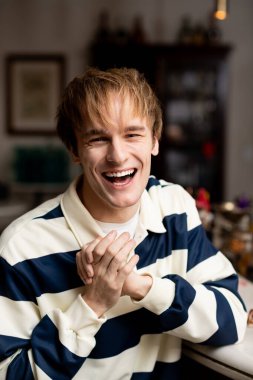 Sarı saçlı neşeli bir genç adam şık bir kafede otururken gülüyor..