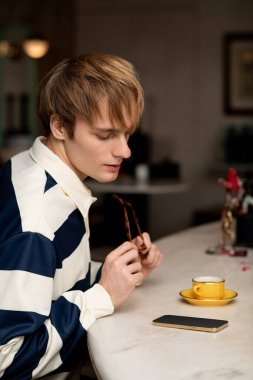 Gündelik giysiler içindeki sarışın genç adam modern bir kafede bir fincan kahvenin tadını çıkarıyor..