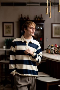 Çağdaş bir kafede, barın yanında dikilen günlük kıyafetli sarışın adam gülümsüyor..