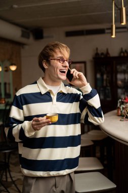 Sarı saçlı genç bir adam telefonda konuşuyor, modern bir kafede elinde fincanla..