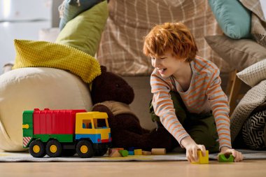 Neşeli çocuk, yumuşak yastıklar ve renkli bloklarla çevrili enerjik bir oyuncak kamyonla oynuyor..