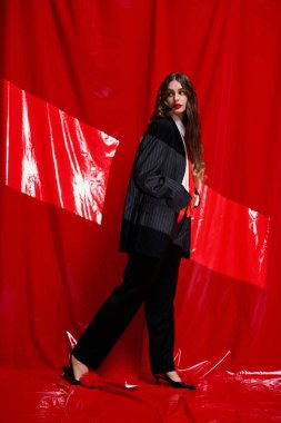 Şık giyinmiş şık bir kadın, koyu kırmızı bir zemine karşı güvenle yürür..