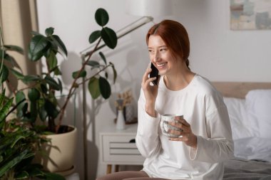 Kızıl saçlı neşeli genç bir kadın modern dairesinde oturur, kahvesini yudumlar ve konuşur..