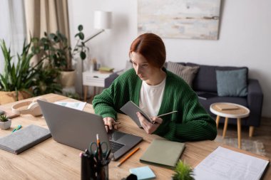 Kızıl saçlı genç bir kadın şık bir ev ortamında dizüstü bilgisayarına odaklanıyor..