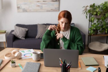 Kızıl saçlı kadın rahat bir apartman dairesinde dizüstü bilgisayarıyla meşgul oluyor..