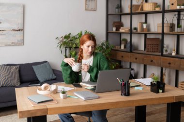 Kızıl saçlı genç bir kadın, modern dairesinde bilgisayarını kullanırken kahvesini yudumluyor..