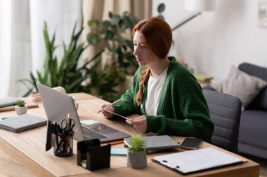Odaklanmış kızıl saçlı kadın, bitkilerle çevrili, modern, rahat bir dairede dizüstü bilgisayarıyla çalışıyor..