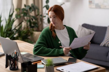 Kızıl saçlı genç bir kadın evindeki rahat bir masada notlarını dikkatle okuyor..